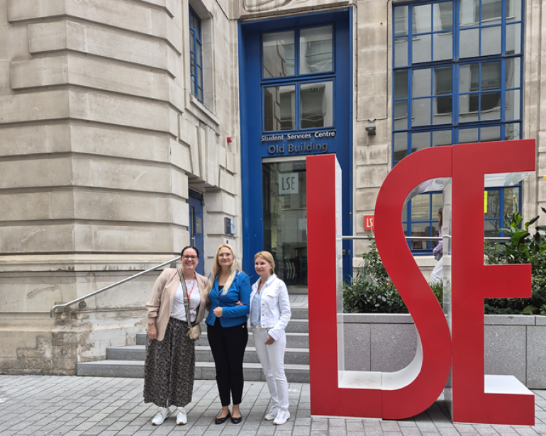 zdjęcie trzech kobiet przed siedzibą London School of Economics, kobiety stoją przy dużym akronimie LSE 
