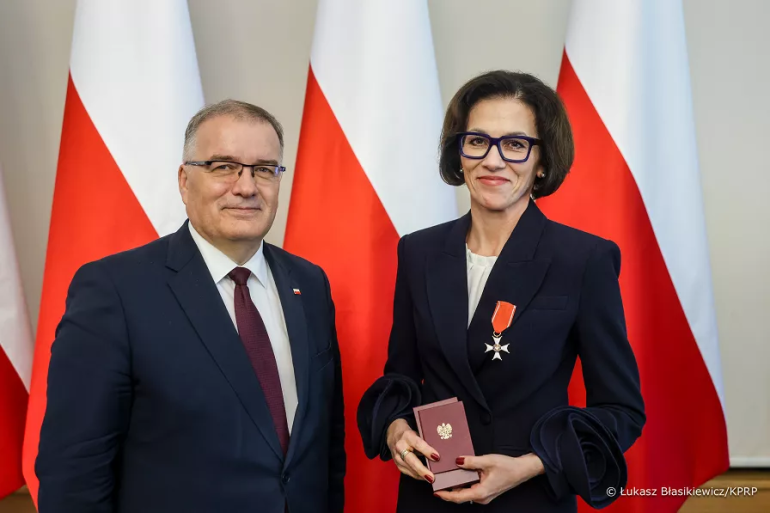 kobieta przyjmuje odznaczenie; zdjęcie na tle trzech polskich flag