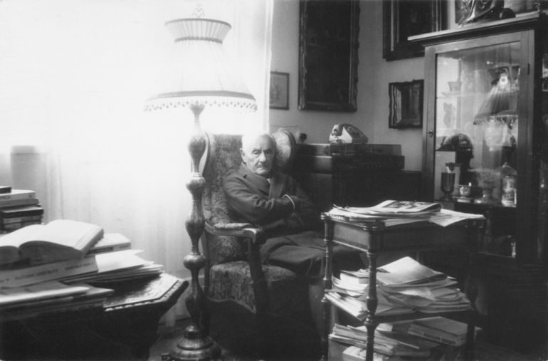 czarno-białe zdjęcie; prof. Lipiński siedzi na fotelu wśród książek i antyków 