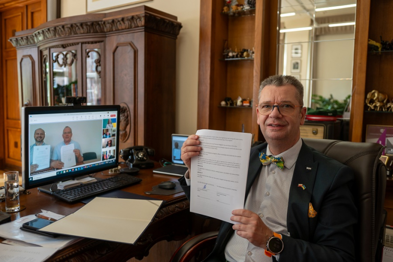 Mężczyzna przy komputerze z podpisaną umową; w tle na ekranie widać dwóch mężczyzn trzymających tę samą umowę w rękach