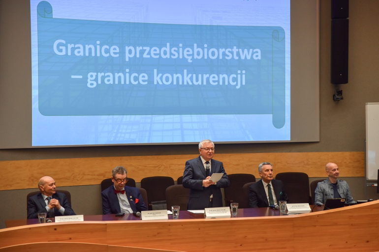 kilku mężczyzn na konferencji naukowej siedzi za stołem prezydialnym, jeden stoi i przemawia