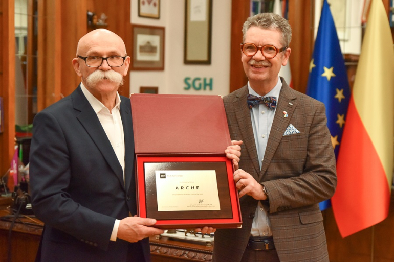 dwóch mężczyzn trzyma w rękach mosiężną tabliczkę informującą o przyjęciu firmy Arche do Klubu Partnerów SGH