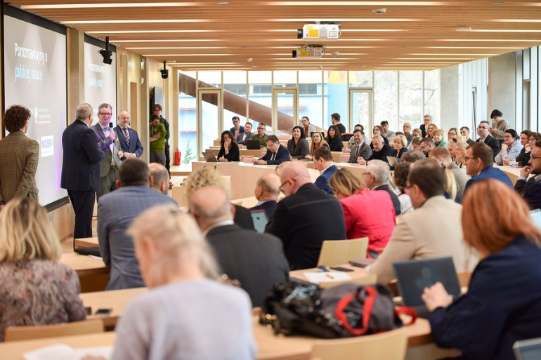 trzech mężczyzn stoi i przemawia w sali konferencyjnej wypełnionej ludźmi