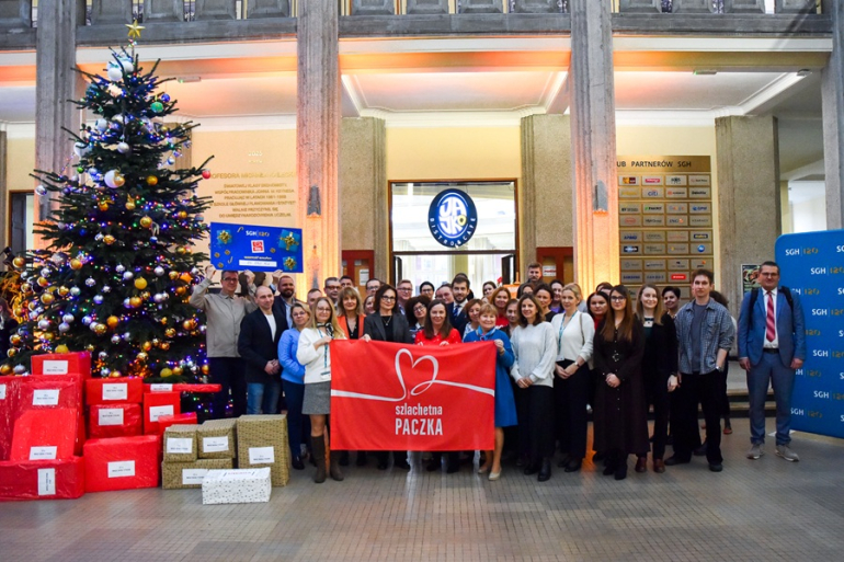kilkanaście osób pozuje do zdjęcia przy dużej choince i prezentach 