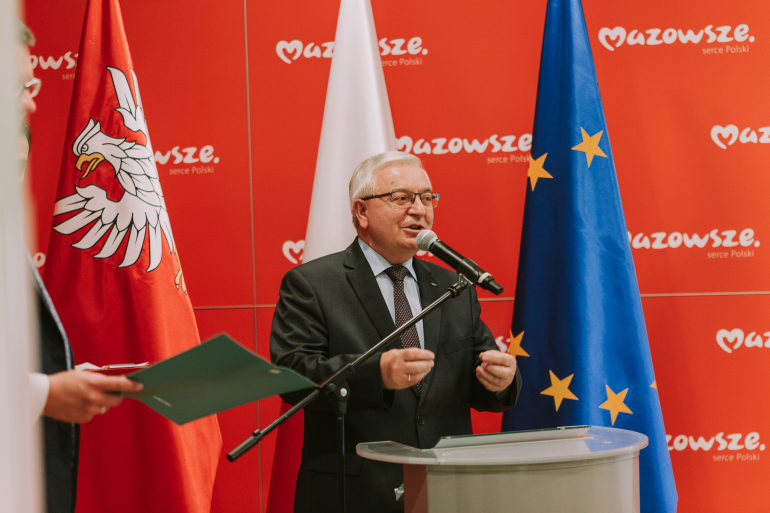 mężczyzna stojący przy mównicy na tle flagi polskiej i unijnej