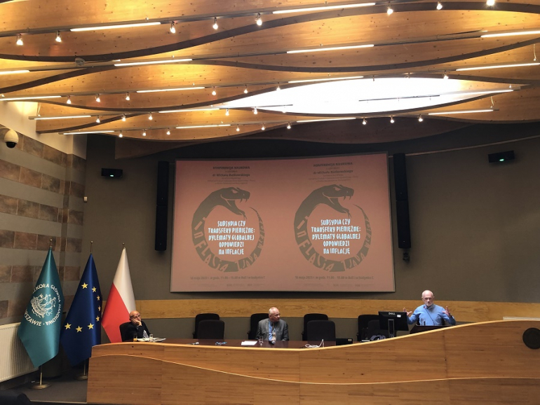 męzczyzna zabiera głos na konferencji w auli, po lewej stronie flaga polski, UE i SGH, za nim na ekranie wyświetlana prezentacja