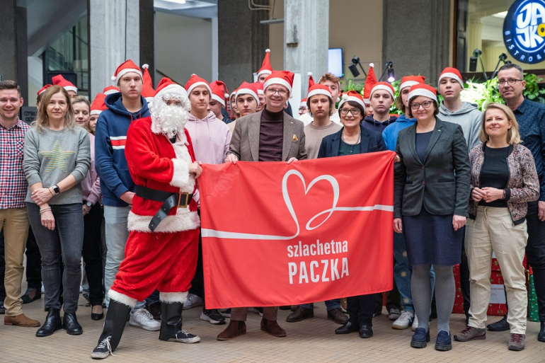 grupa osób w czapkach mikołajowych stoi przed ubraną choinką; częśc z nich w rękach trzyma czerwony transparent z napisem Szlachetna Paczka