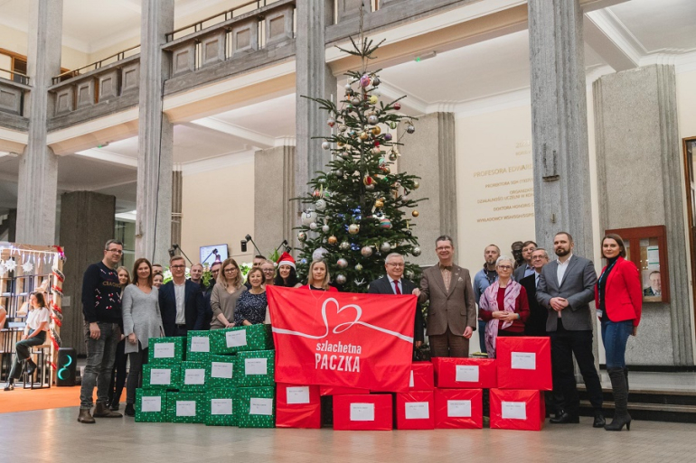 grupa osób stoi przed choinką i kilkunastoma paczkami zebranymi w ramach akcji Szlachetna Paczka; w ręku trzymają banner z napisem: Szlachetna Paczka