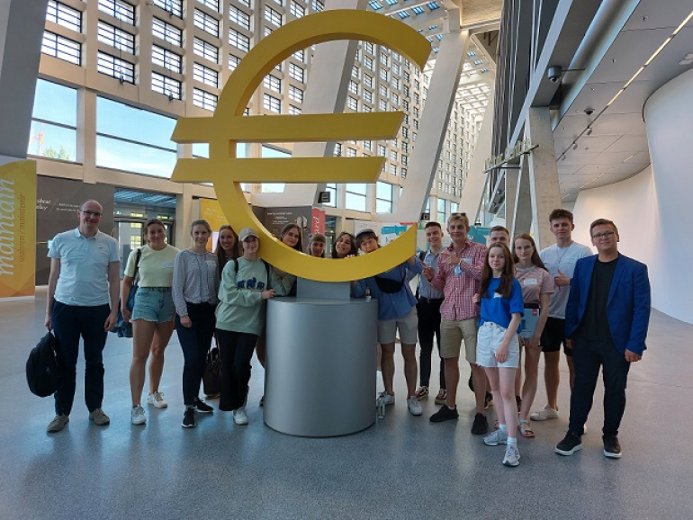 grupa młodzieży stoi przy olbrzymim symbolu euro w siedzibie EBC