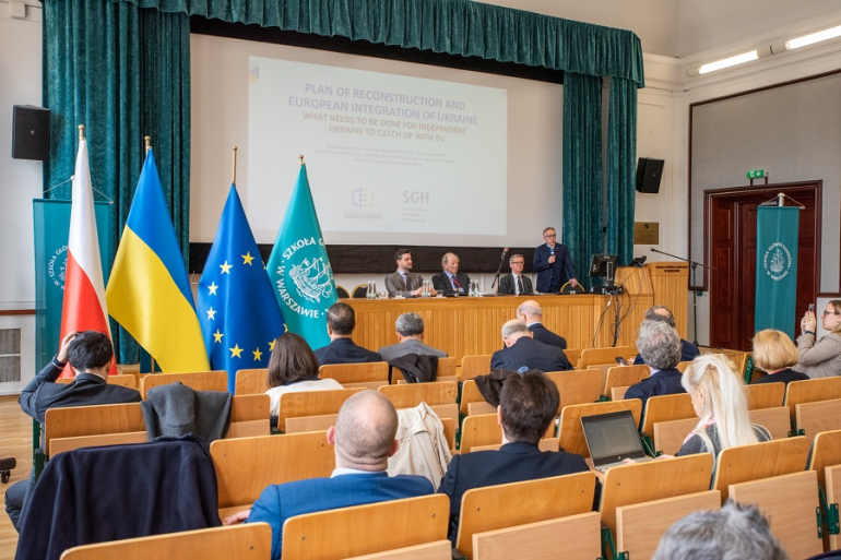 grupa osób siedzi na sali, na scenie kilka osób siedzi za stołem prezydialnym; po lewej stronie sceny flagi Polski, Ukrainy, UE, SGH
