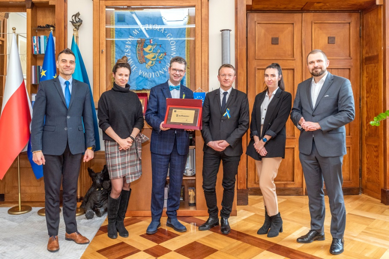 grupa osób stojąca w gabinecie; w tle flaga RP, UE i sztandar SGH