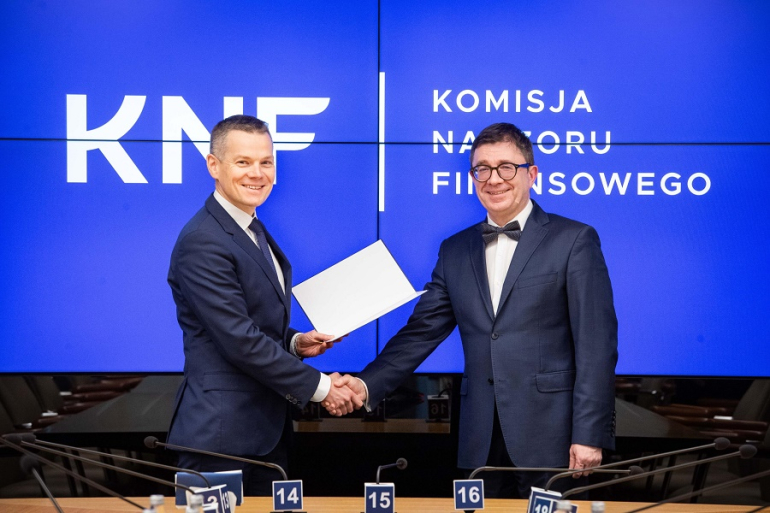 mężczyzna przekazuje dyplom drugiemu mężczyźnie; w tle niebieskie tło z logiem i nazwą KNF